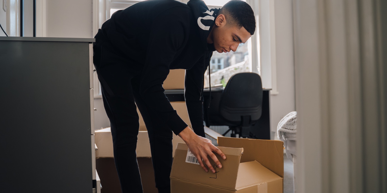 A young guy in a black hoodie and jogger packs stuff into a cardboard box in his bedroom - he's super prepared because he read the fused student moving guide