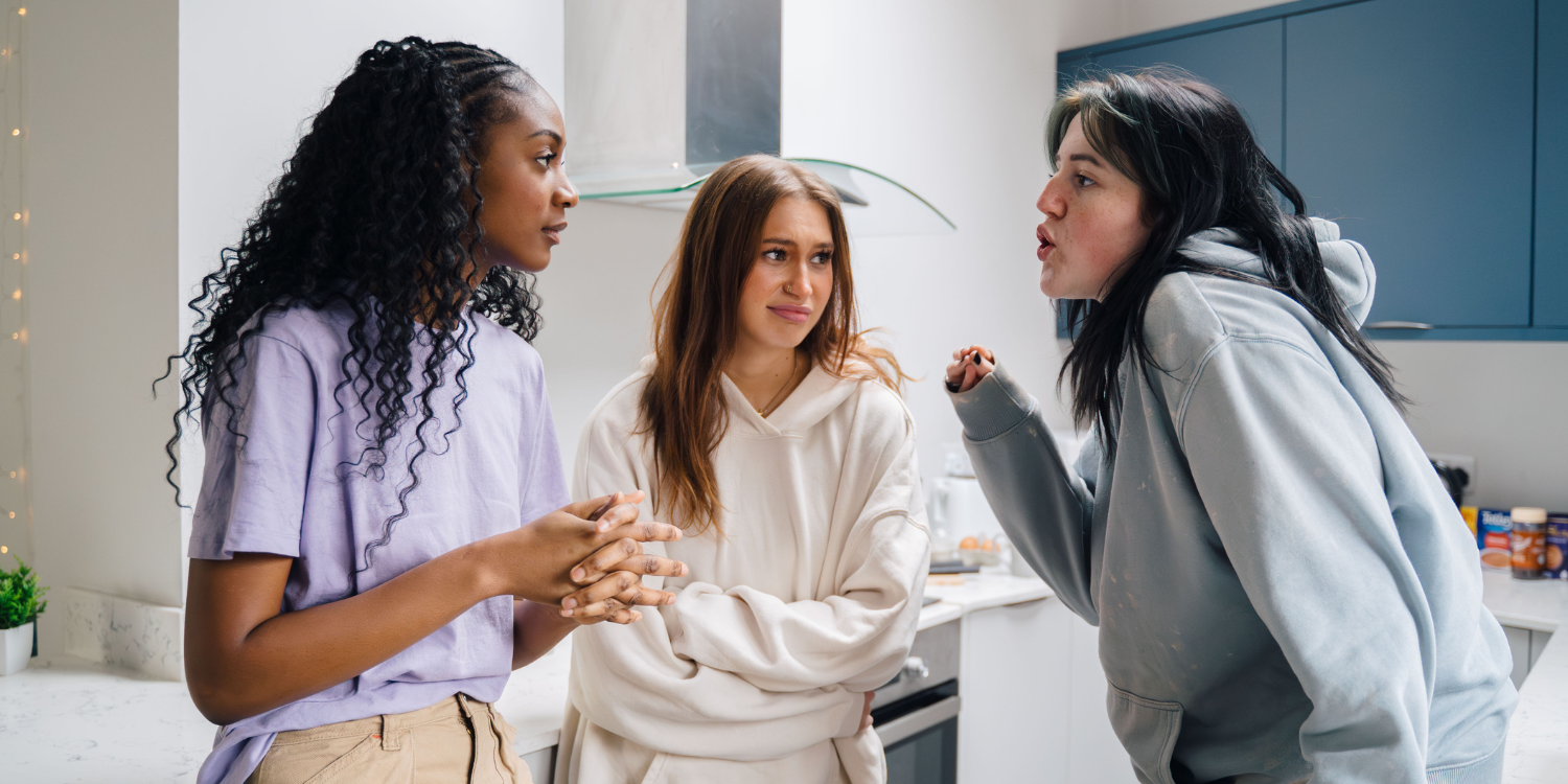 Three young women stand in a kitchen facing the camera - obviously arguing about student bills - two of them are looking at the other, who is pointing and looks annoyed. They seem unimpressed.