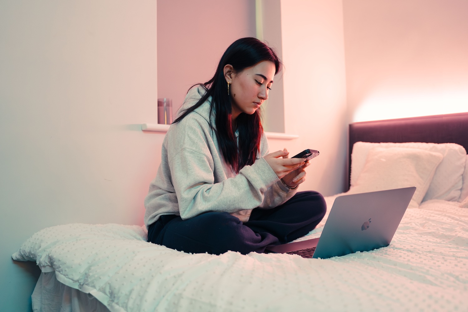 A young asian woman sits on a bed cross-legged in cosy joggers and a white baggy hoody. She has her laptop in front of her and is scrolling on her phone