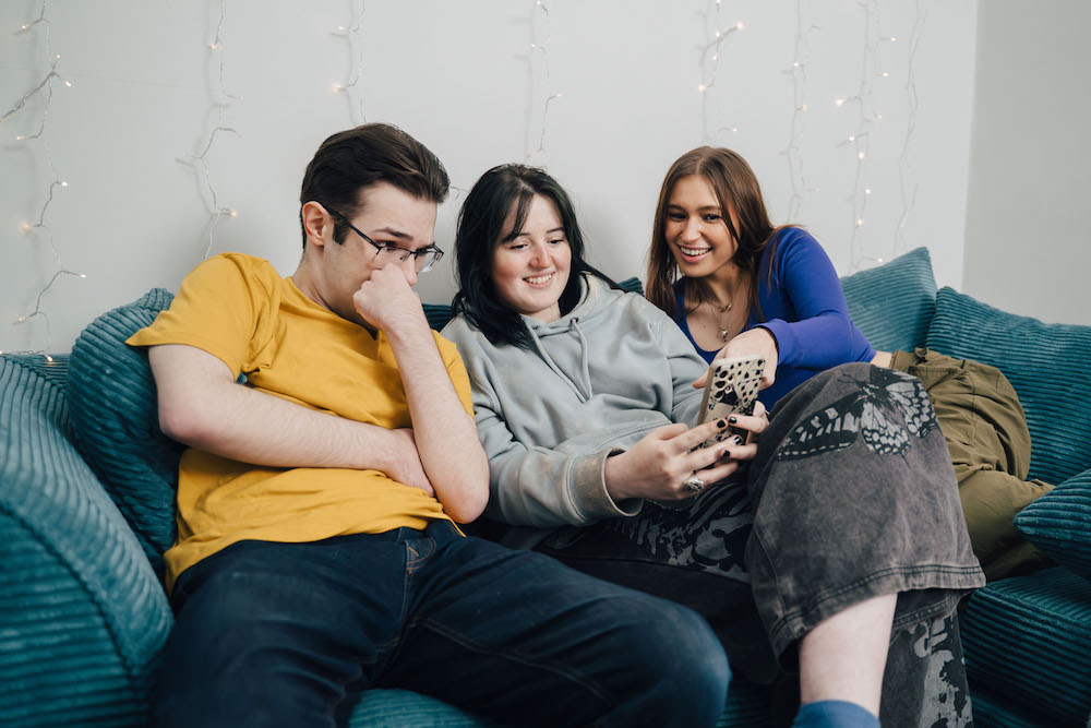 Three students sit on a sofa looking at the middle student's phone deciding if a student bills package is the best choice for them