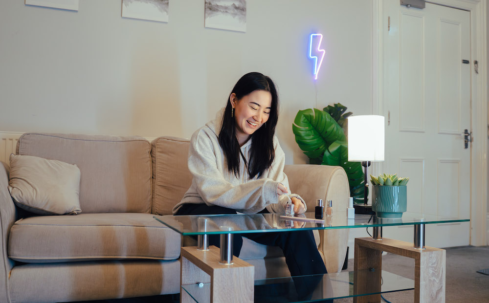 A young student sits painting her nails on the sofa. A lightning bolt neon sign floats behind her. She's very relaxed because she has Unlimited Energy in her Fused Student Bills package